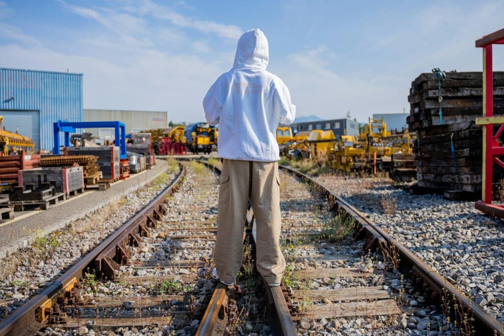 A person in a white hoodie stands on railway tracks in an industrial area on a sunny day.