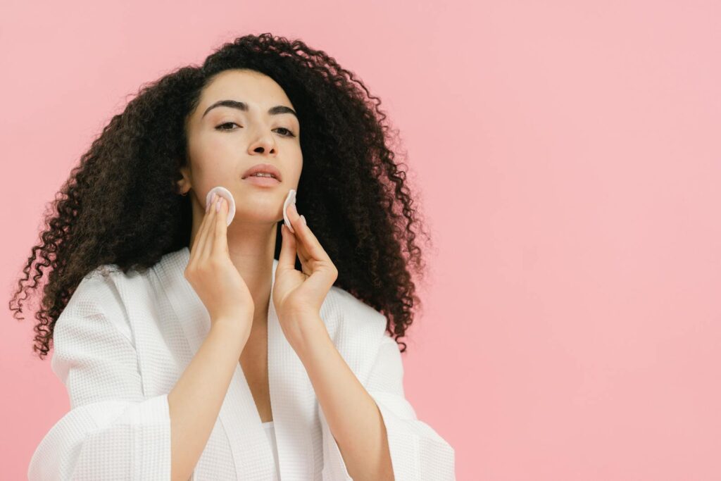 A young woman with curly hair using cotton pads for her skincare routine against a pink background.