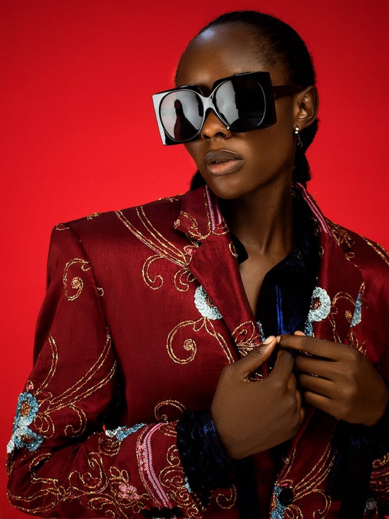 Fashionable African woman posing in embroidered blazer and oversized sunglasses against red backdrop.