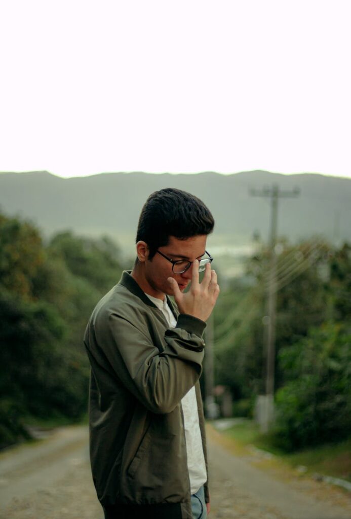 Man wearing glasses and bomber jacket standing outdoors with blurred greenery.