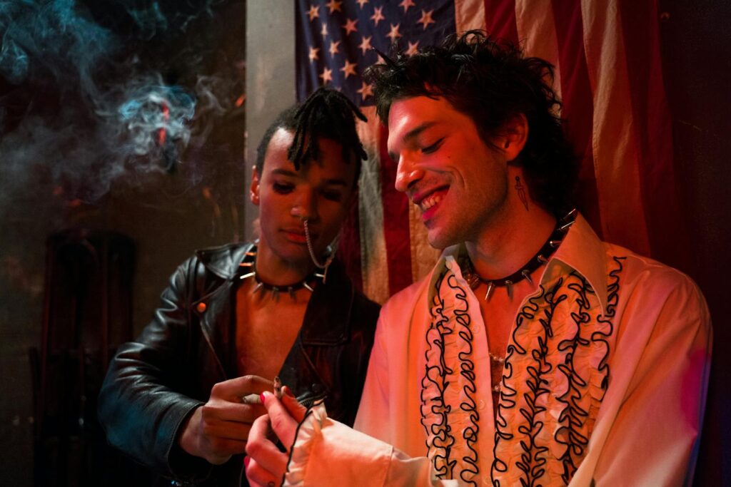 Two stylish men in punk outfits enjoying a smoke indoors with an American flag backdrop.