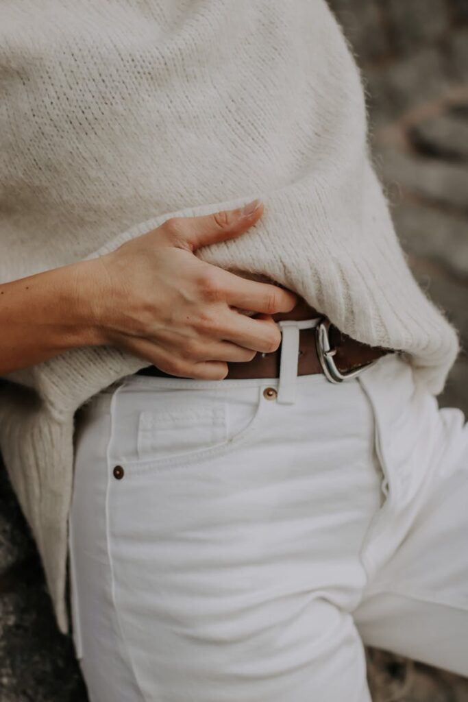 Hand adjusting belt on white trousers with cozy sweater, showcasing casual fashion style.