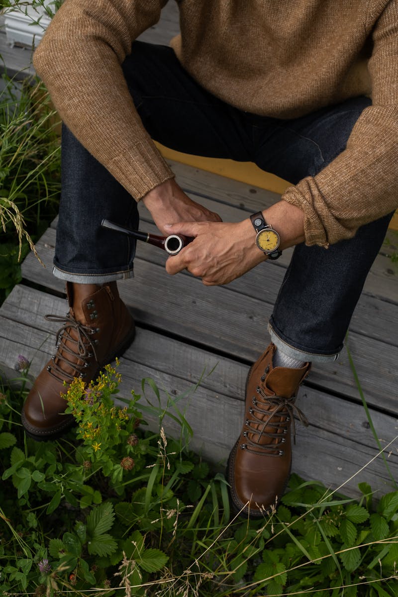 Senior man sitting on a porch, wearing a sweater and boots, holding a pipe.