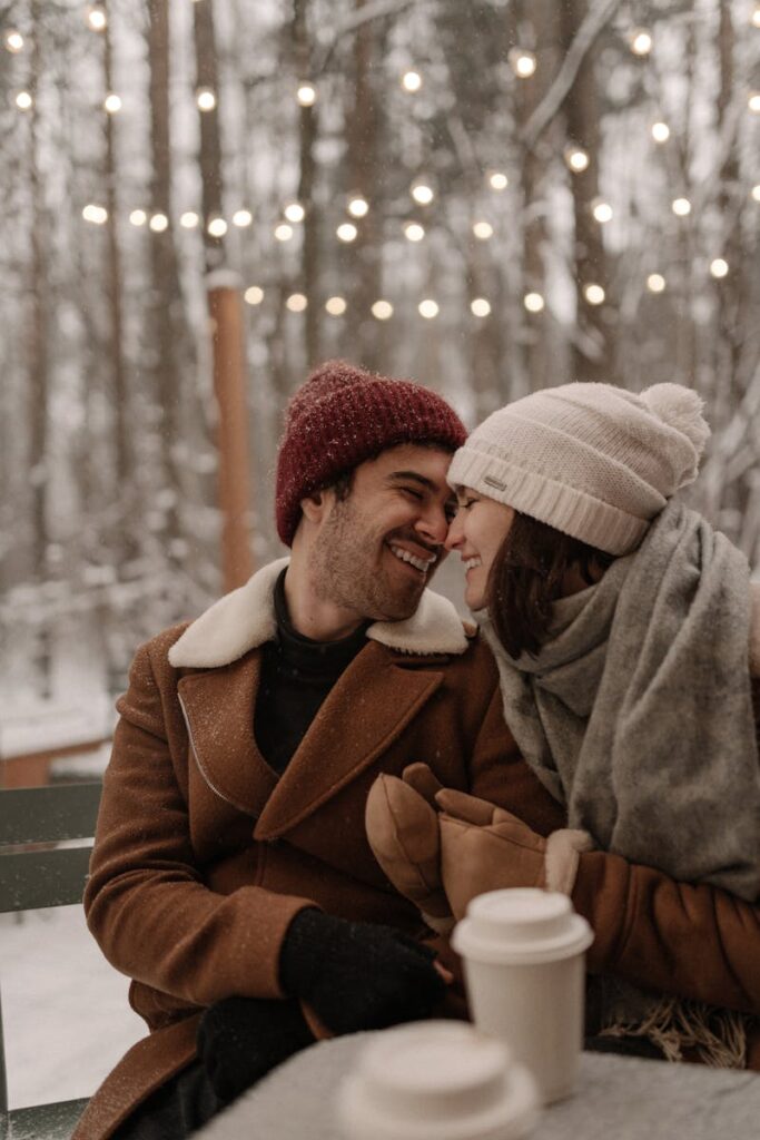 A joyful couple warmly dressed, embracing in a snowy forest with hot drinks, under string lights.