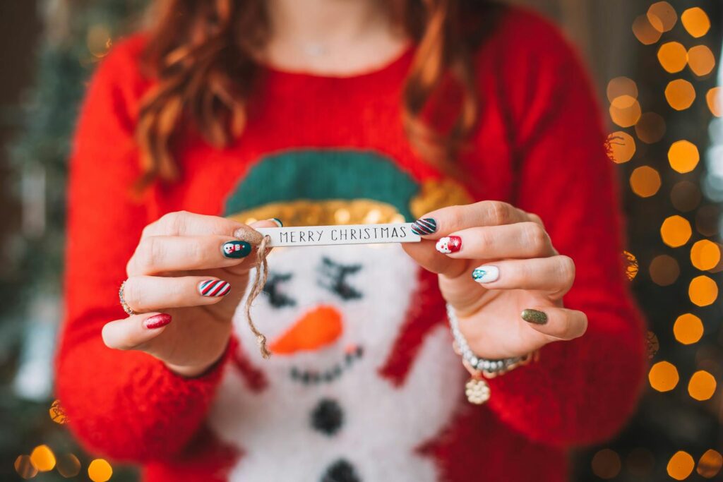 Close-up of hands holding a Merry Christmas ribbon, wearing a festive red sweater with cheerful nail art.