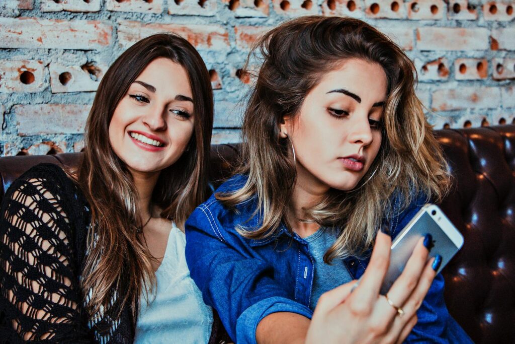 Two young women capturing a selfie while sitting in a casual indoor setting, smiling and relaxed.