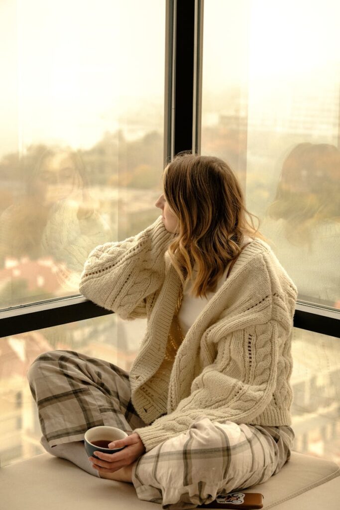Woman enjoying a quiet moment with a hot drink, gazing through a window, dressed warmly.