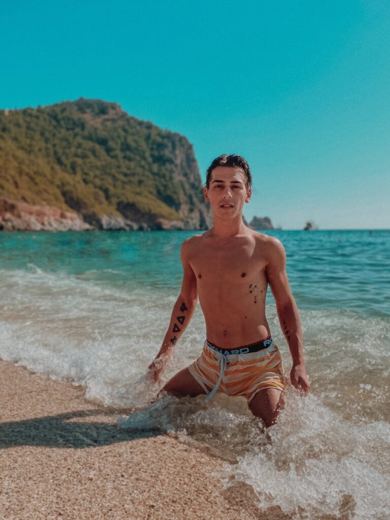 Shirtless young man kneeling at the beach in Alanya, Turkey, with a scenic backdrop.