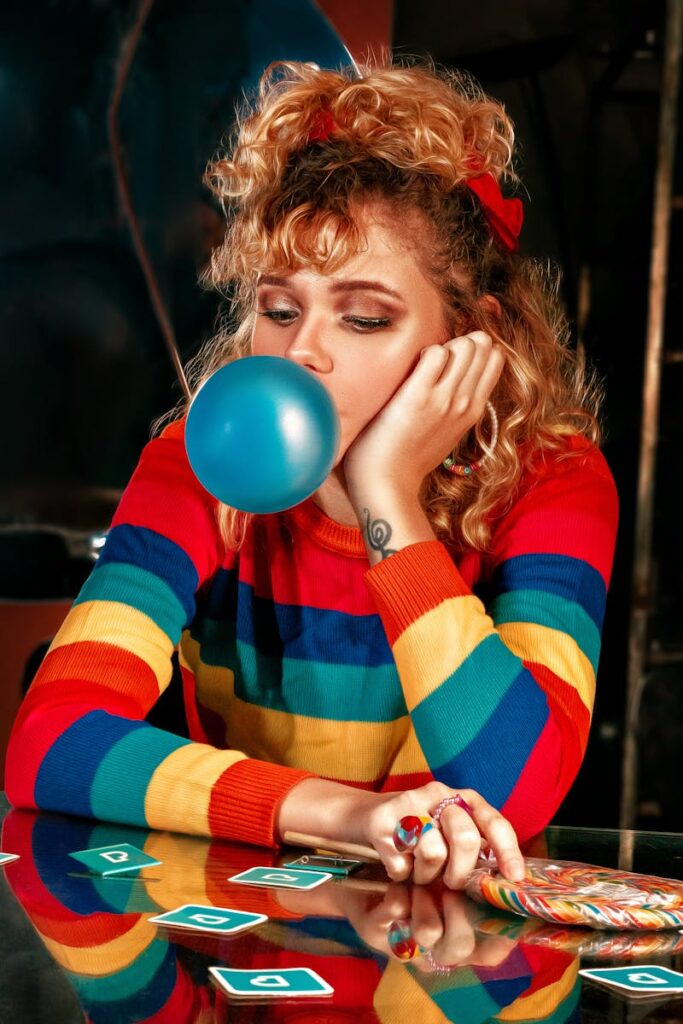 Woman in a rainbow sweater blowing bubblegum in a retro scene indoors, conveying a playful and nostalgic vibe.