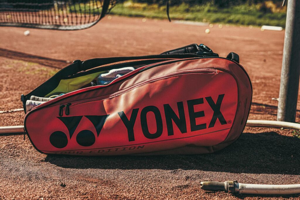Close-up of a Yonex tennis bag lying on a clay court under sunlight, showcasing sports gear.