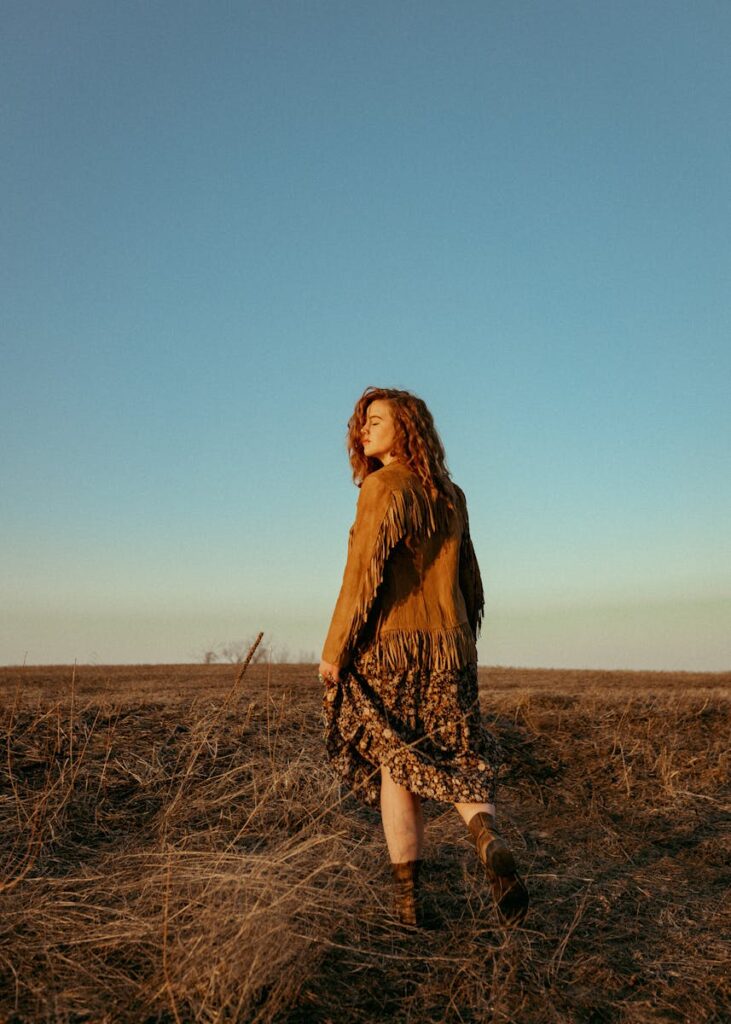 A stylish woman in a fringe jacket walks through dry fields in IA, USA during sunset.