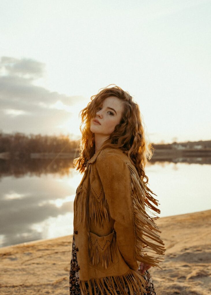 A captivating portrait of a woman in a fringed jacket by a serene river at sunset, capturing natural beauty and tranquility.