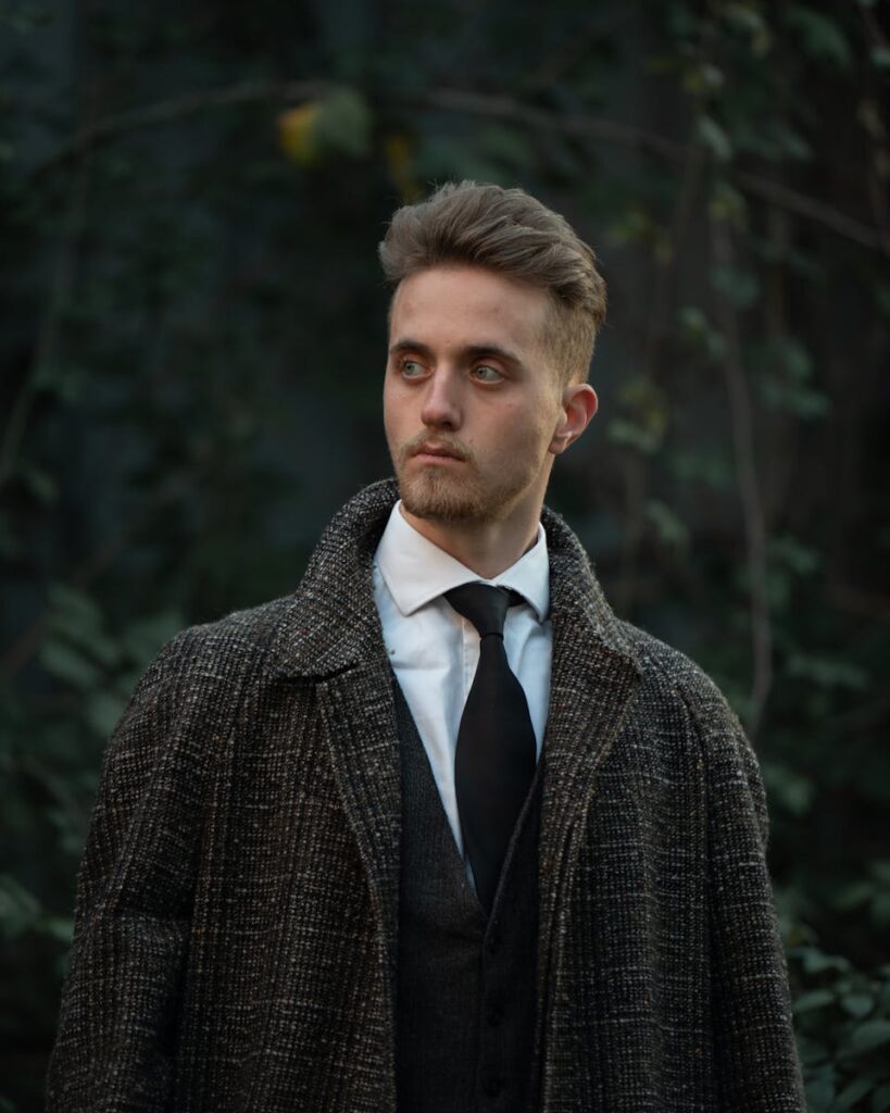 Man in formal attire and tweed coat in an outdoor setting with lush greenery.