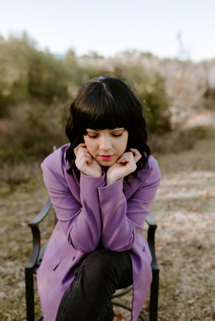 Thoughtful woman with black hair in a purple coat sitting outdoors on a chair.
