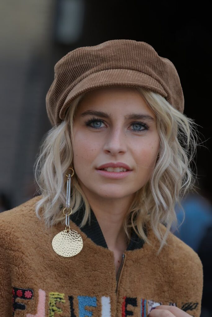 Portrait of a stylish woman in a hat with blonde hair and a brown jacket smiling outdoors.