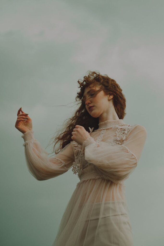 Red-haired woman in a bohemian dress poses outdoors against an overcast sky.