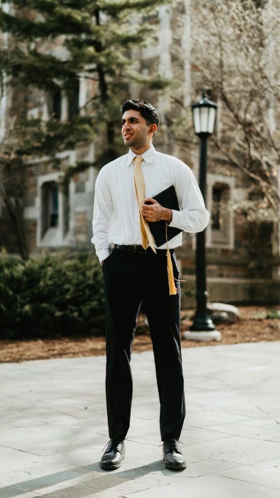 Young man standing outdoors in graduation attire holding his cap, showcasing success.