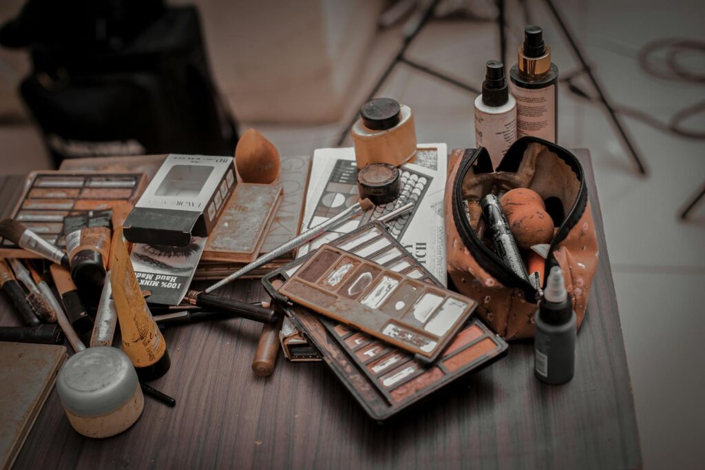 A collection of used makeup products, including brushes and palettes, on a wooden table indoors.