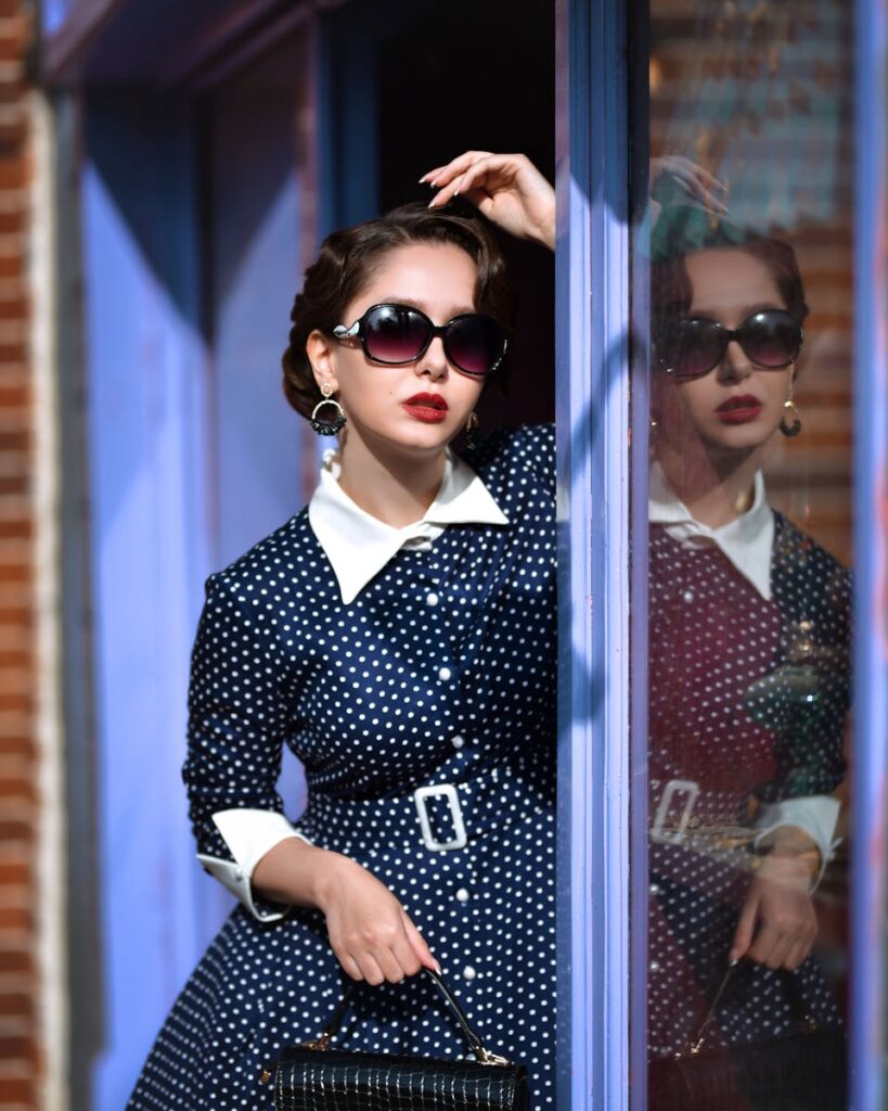 Elegant woman in a vintage polka dot dress reflecting in shop window, exuding retro fashion style.