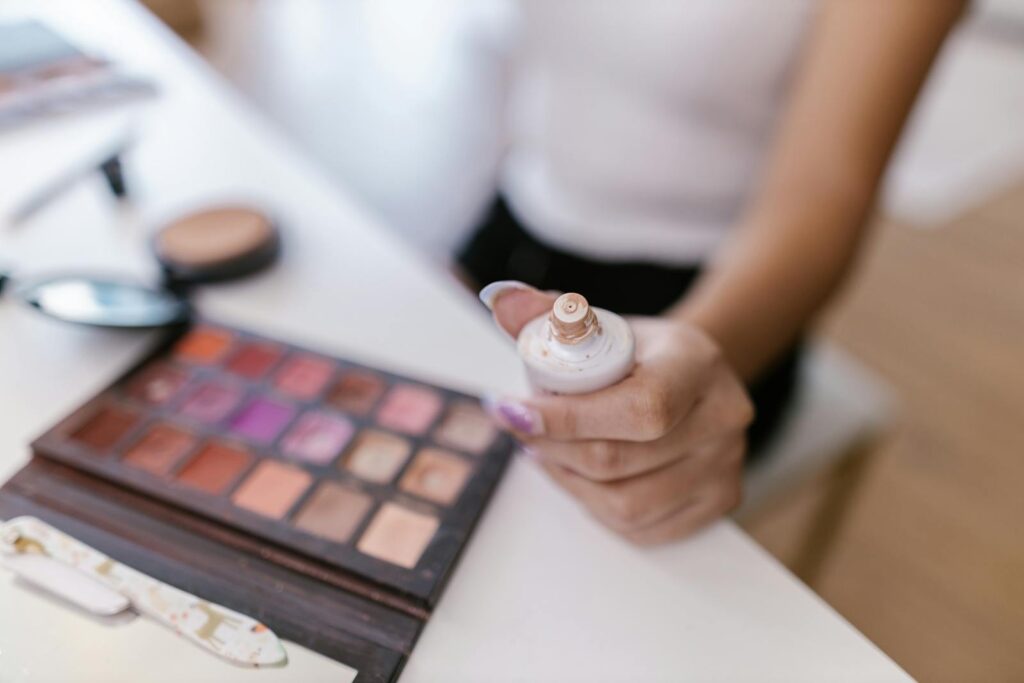 Close-up of a person holding foundation bottle with colorful makeup palette in focus.
