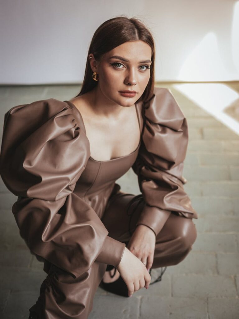Fashion portrait of a woman in a brown leather outfit with puffed sleeves, posing indoors.