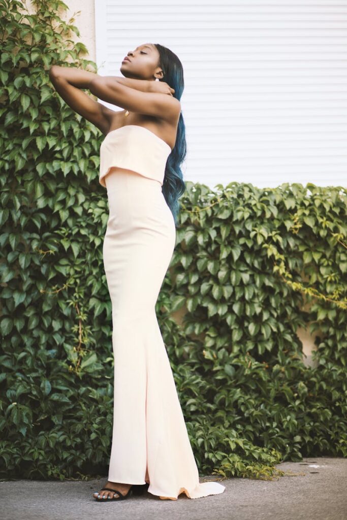A stylish black woman in a strapless gown poses in front of lush greenery.