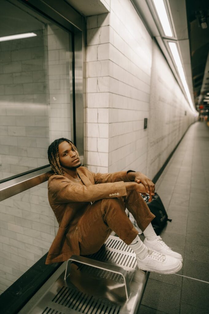 Stylish man in brown suit with braided hair sitting on platform bench in subway station.