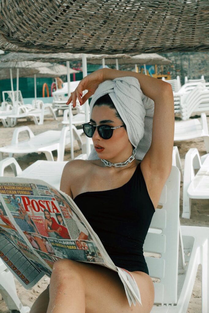 Stylish woman in swimsuit reading a newspaper with towel on head at beach.