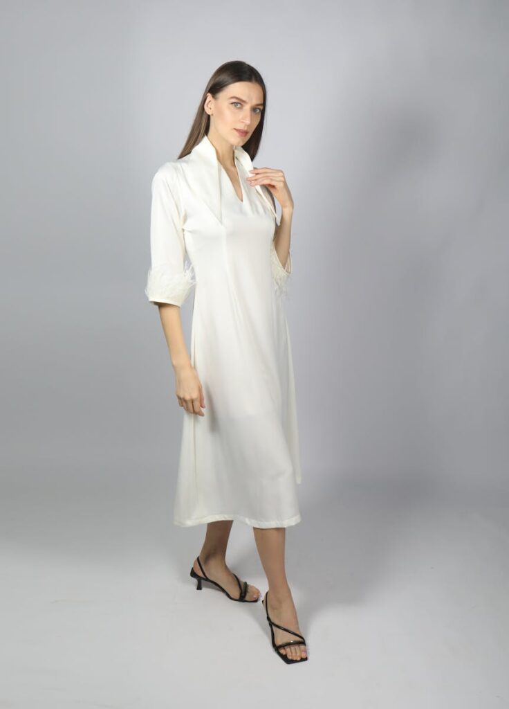 Fashionable woman in a simple white dress posing gracefully indoors under studio lighting.