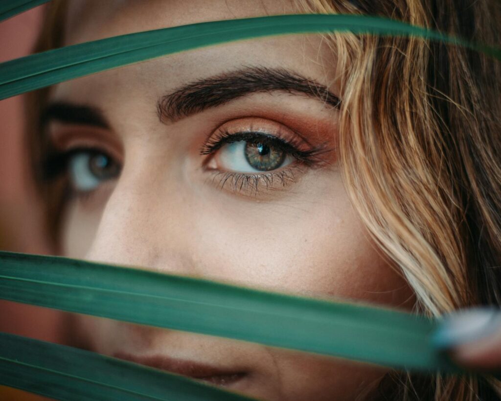 A close-up portrait of a woman's eye peeking through green leaves, evoking mystery and nature.