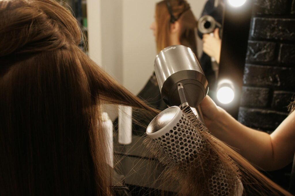 A stylist blow-dries a woman's hair using a brush and hairdryer in a salon.