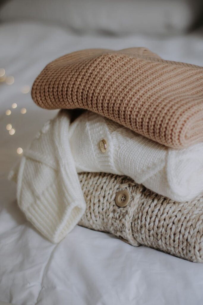 A neatly stacked pile of cozy knitted sweaters in neutral tones on a bed.