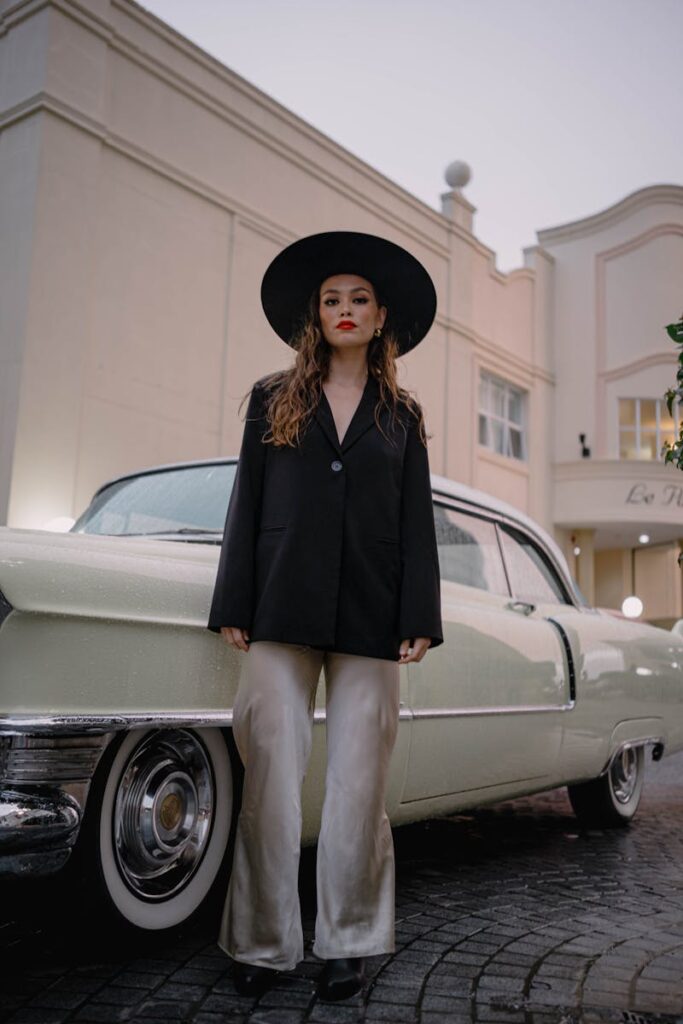 A fashionable woman with a hat stands in front of a vintage car, exuding elegance.