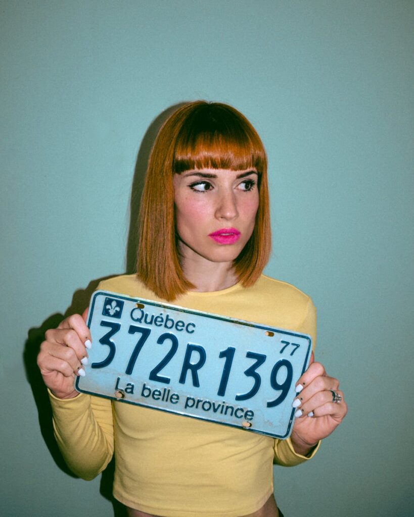 Stylish woman with red hair holding a Quebec license plate against a turquoise background.