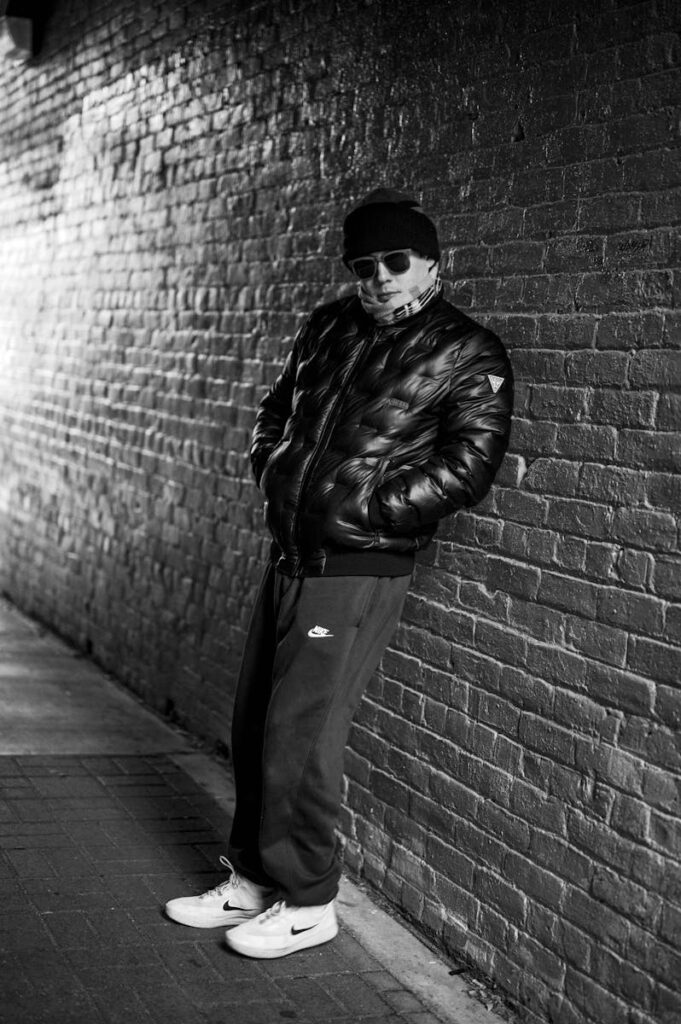 A confident man wearing a jacket poses against a brick wall in black and white.