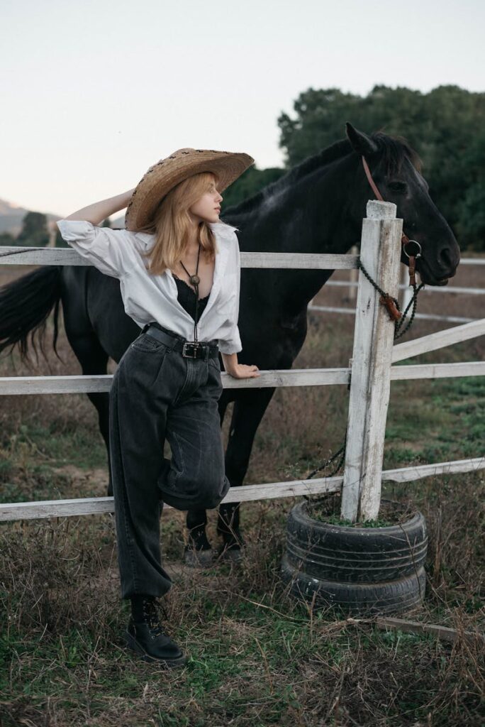 A woman in a cowboy hat leans against a fence with a horse in a rural setting.