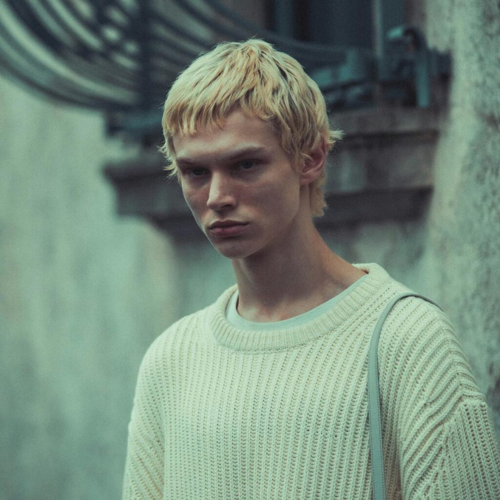 Close-up of a blonde man wearing a knit sweater outdoors with a moody background.