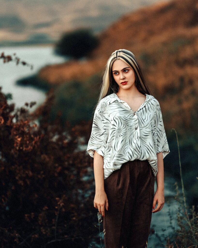 Fashionable woman outdoors by the water at sunset, wearing a patterned blouse and brown pants.