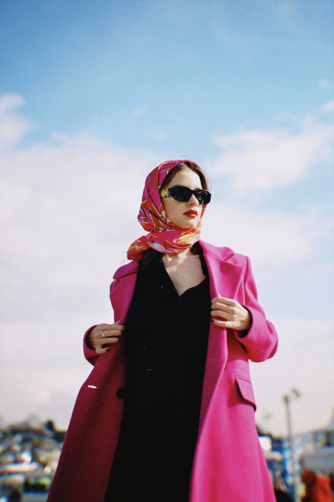 Fashionable woman in a pink coat and scarf, wearing sunglasses, outdoors against a blue sky.