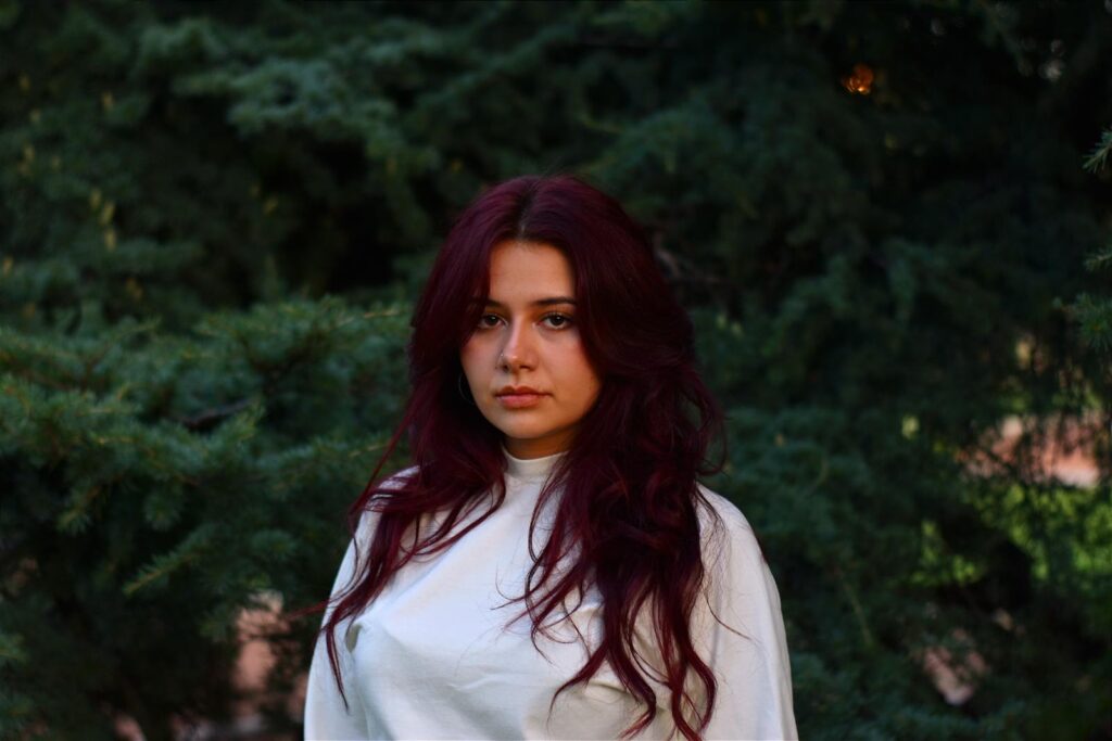 A woman with long burgundy hair poses outside in front of lush greenery in Mendoza, Argentina.