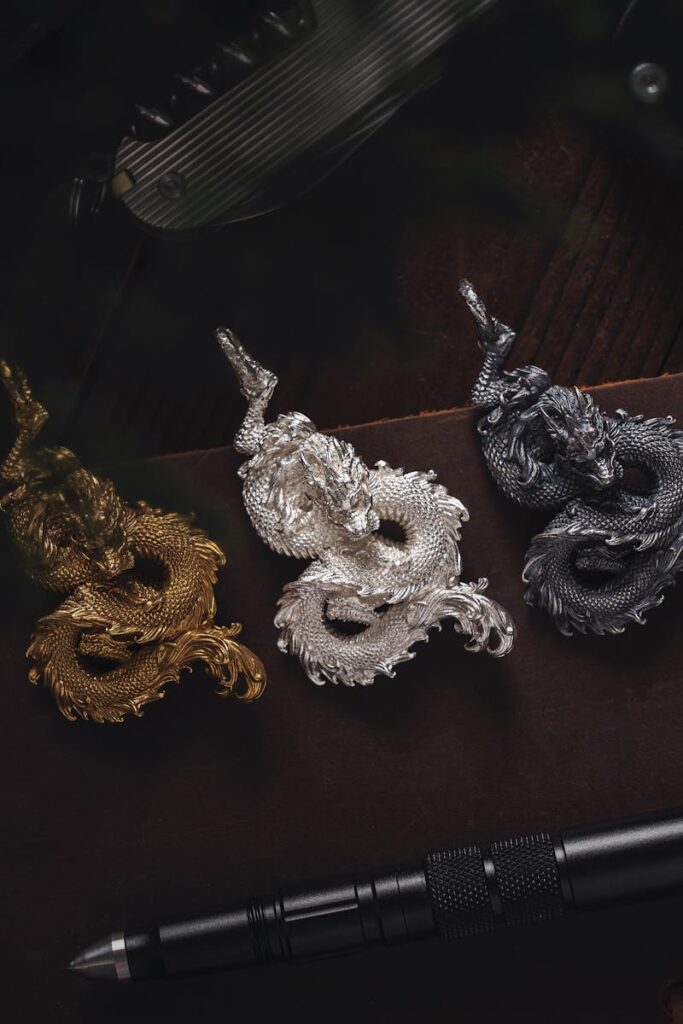 Three intricately designed dragon pendants in gold, silver, and black alongside a tactical pen on a dark surface.