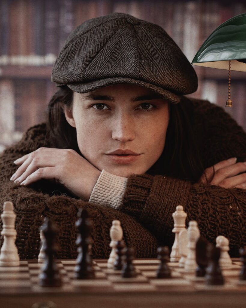 Woman in brown sweater and cap posing behind a chessboard indoors.