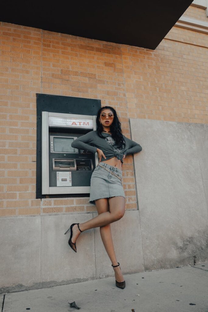 Chic woman in a fashionable outfit posing by an outdoor ATM on a sunny day.