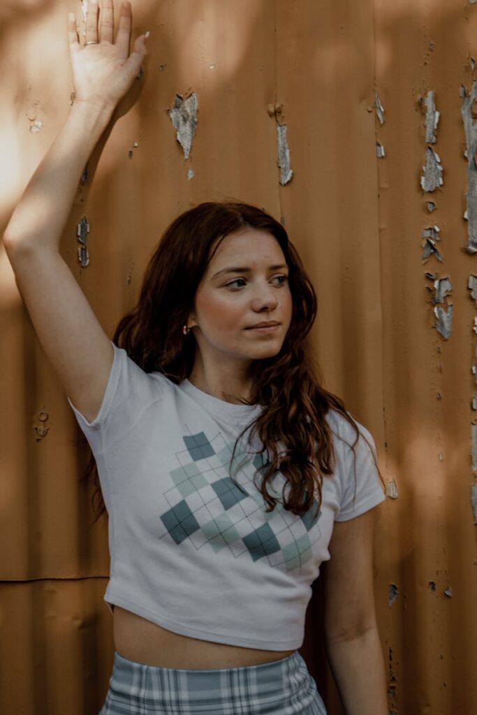 A young woman poses against a textured, rustic wall in natural light, exuding modern fashion vibes.