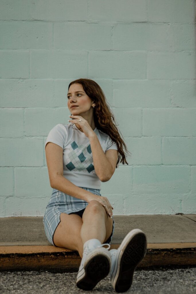 A young woman in casual attire sitting against a pastel wall outdoors.