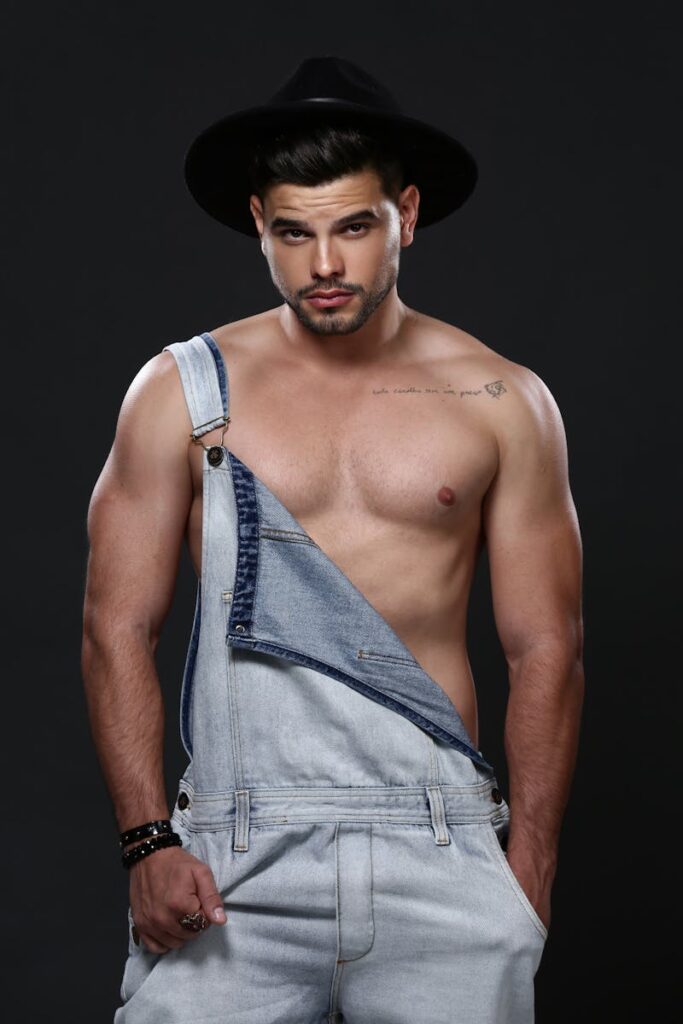 Fashionable man posing confidently in denim overalls with a black hat on a gray studio background.