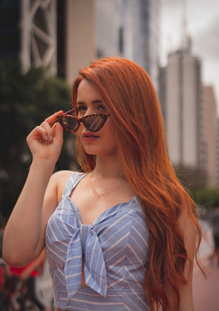 Red-haired woman posing with sunglasses in urban setting, embodying confidence and style.