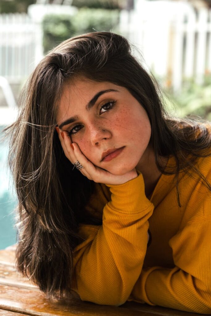 A young woman in a yellow sweater poses thoughtfully by a poolside.