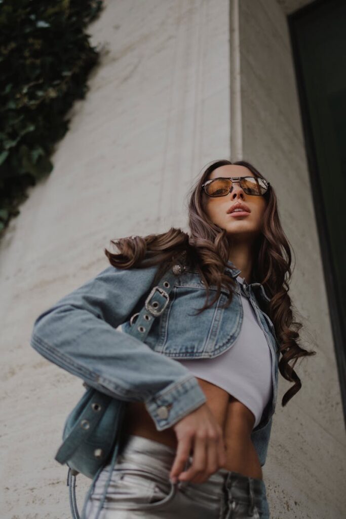 Stylish woman posing in a denim jacket, sunglasses, and crop top with modern vibes.