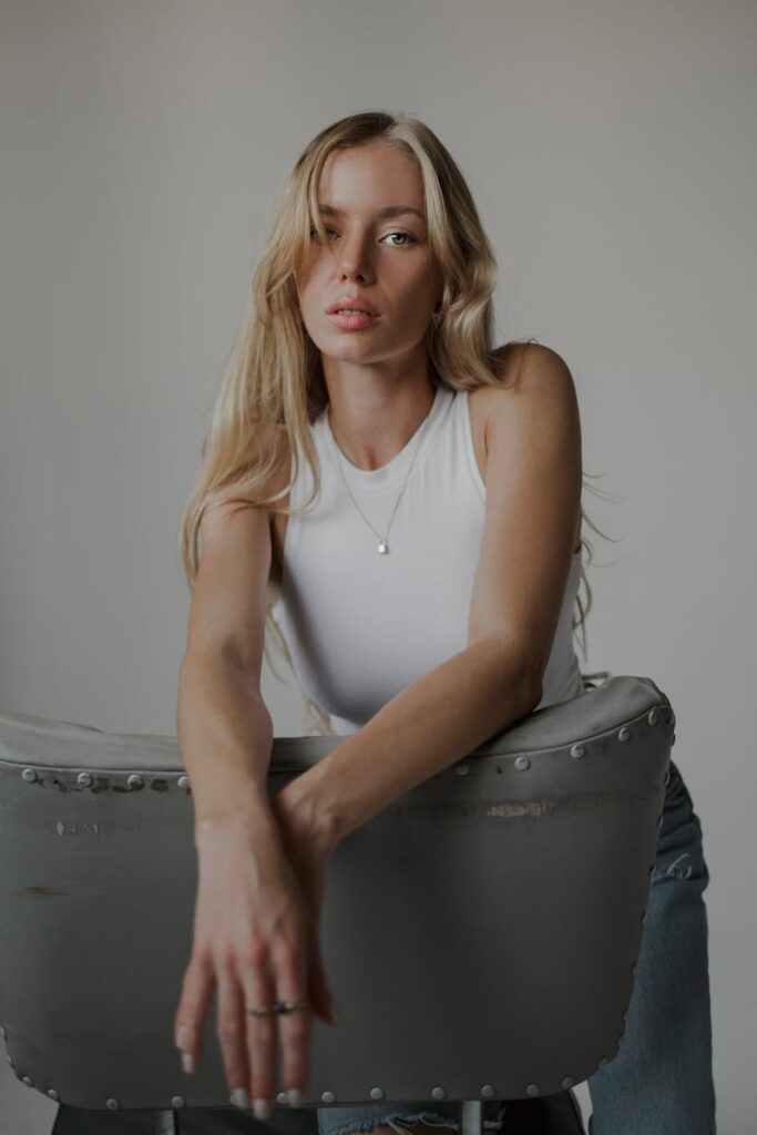 Fashion portrait of a blonde woman in a white tank top posing on a chair indoors. Stylish and modern vibe.